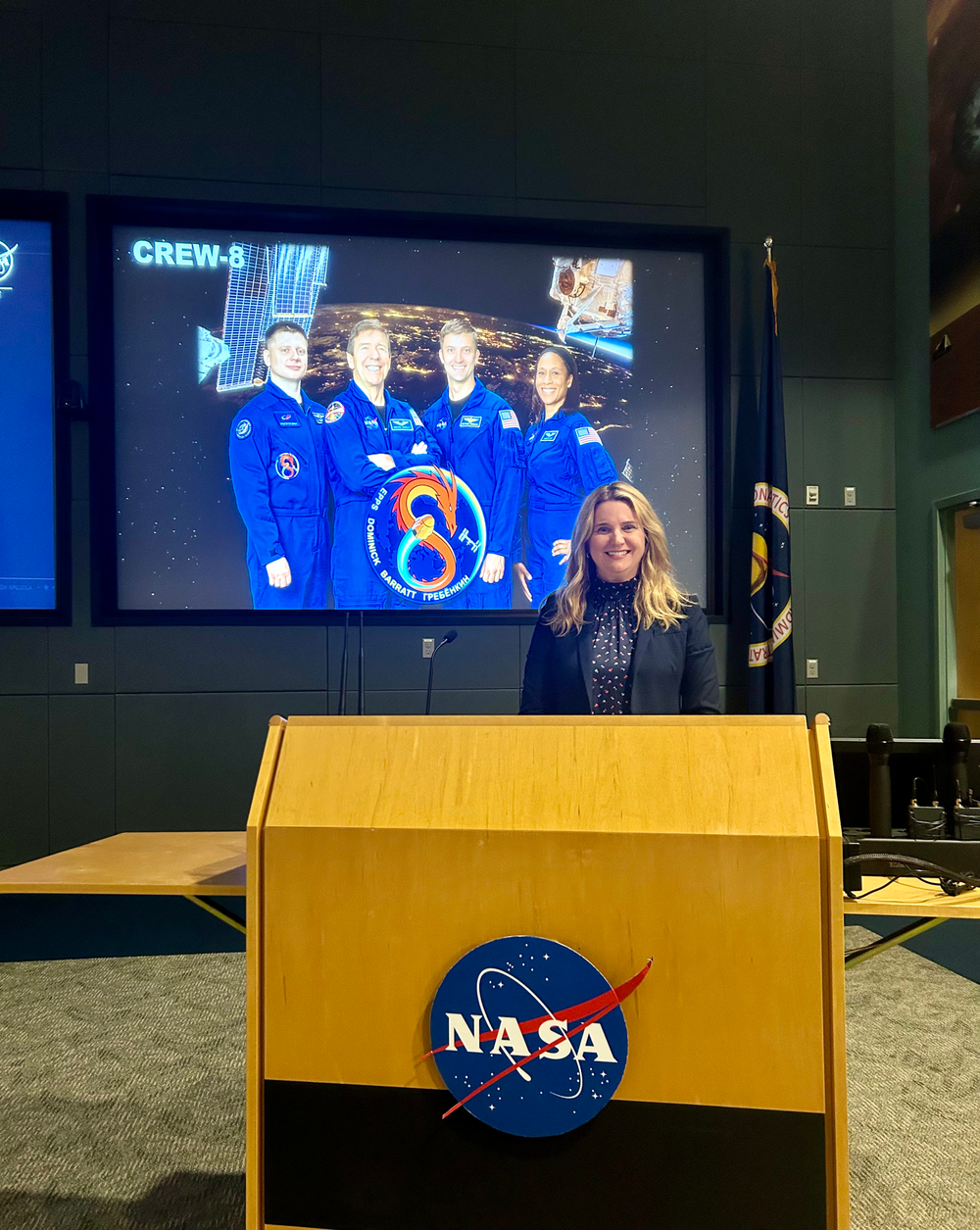 A woman stands behind a podium with a monitor screen and a NASA flag behind her.